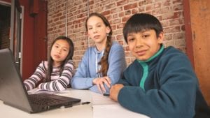 Three children sit at a table and use a laptop.