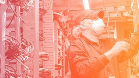 A masked technician attends to a wall of technology equipment, adjusting cables.