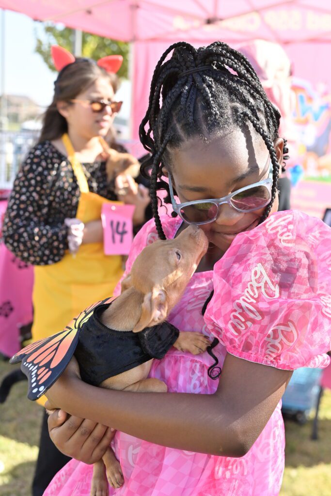 A young girl wearing a pink Barbie dress and sunglasses hugs a small tan dog wearing a butterfly costume at a doggie fashion show benefitting the St. Francis Homeless Project