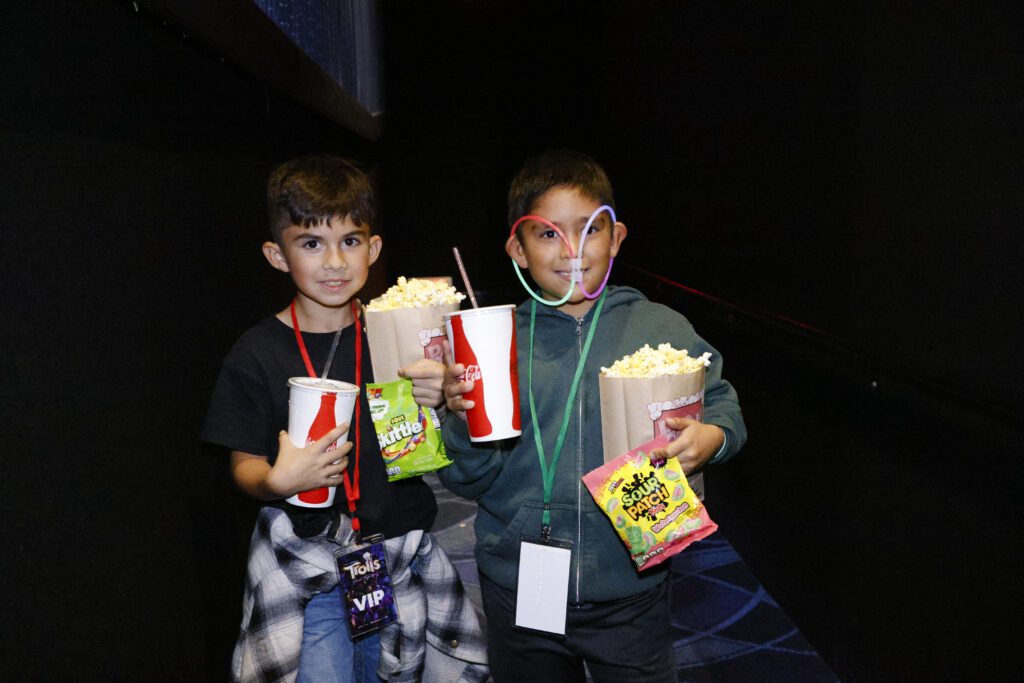 Two young children smile and hold snacks in both hands at the movie theatre.