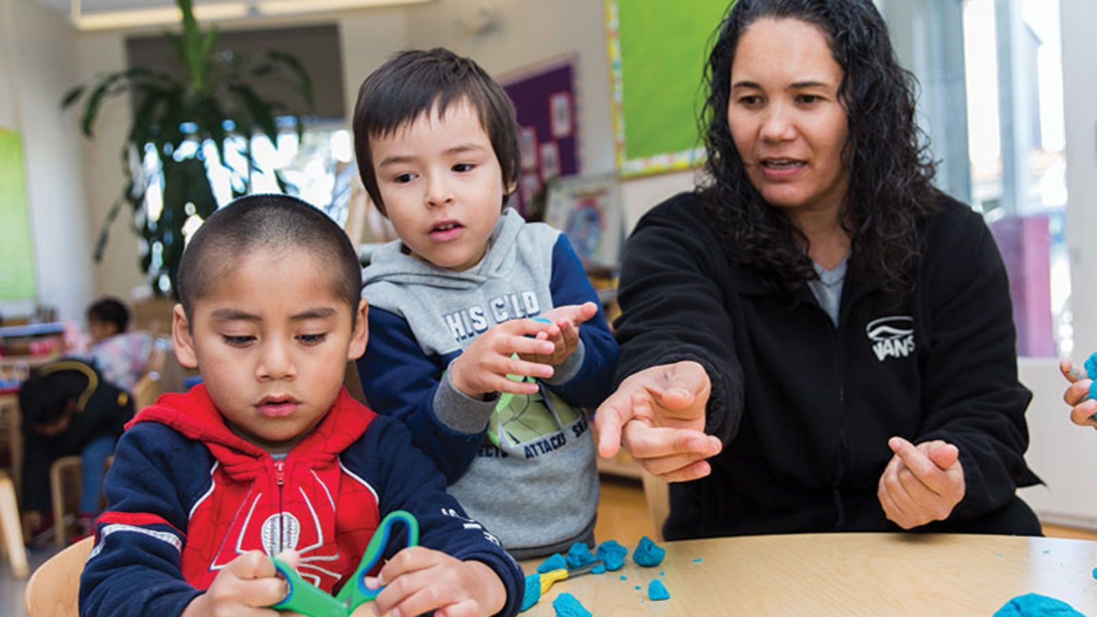 Two children and an adult play with playdough at a table in a classroom.
