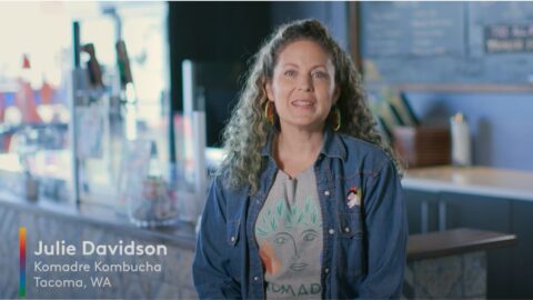 Julia Davidson smiles at the camera with long curly, brown hair, wearing a jean jacket and gray shirt.