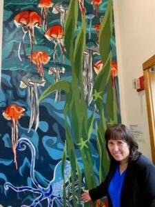 A woman poses with a tall art mural of blue and orange jellyfish underwater.