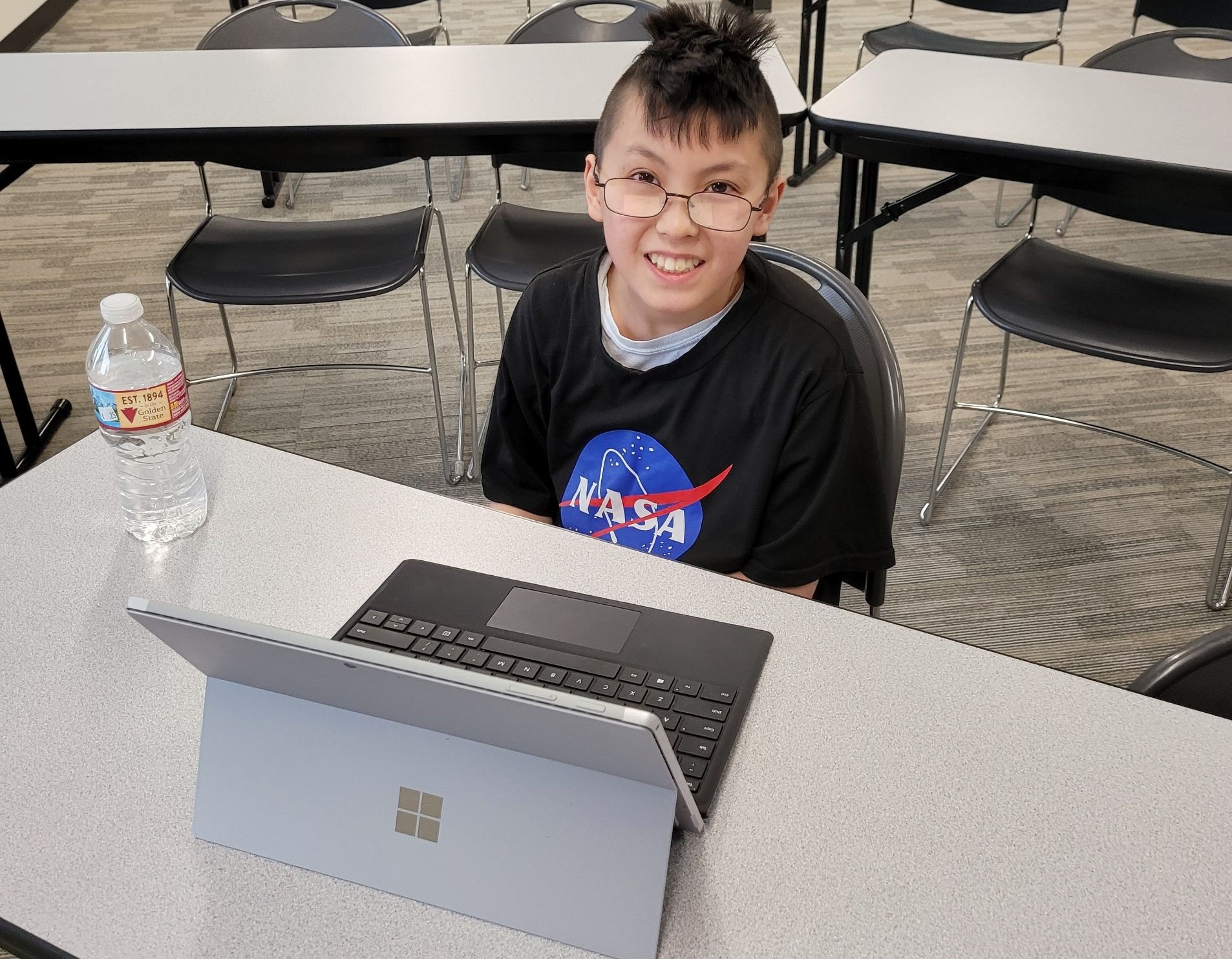 A student wearing a NASA shirt smiles at a desk with a laptop.