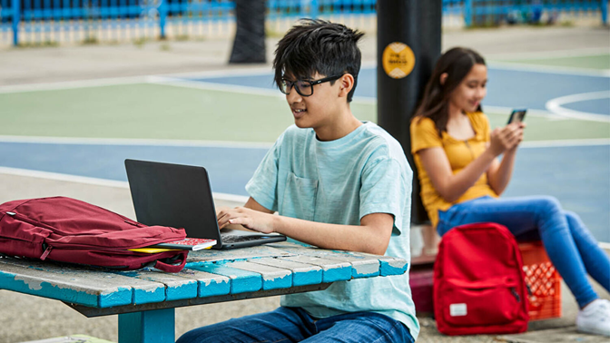 A boy sits at an outdoor table using a laptop computer.