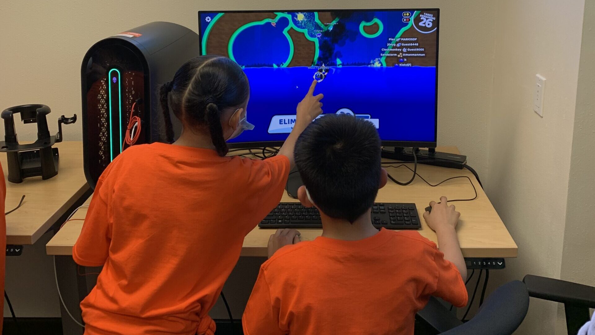 One child uses a mouse on a computer as another child points to the screen.