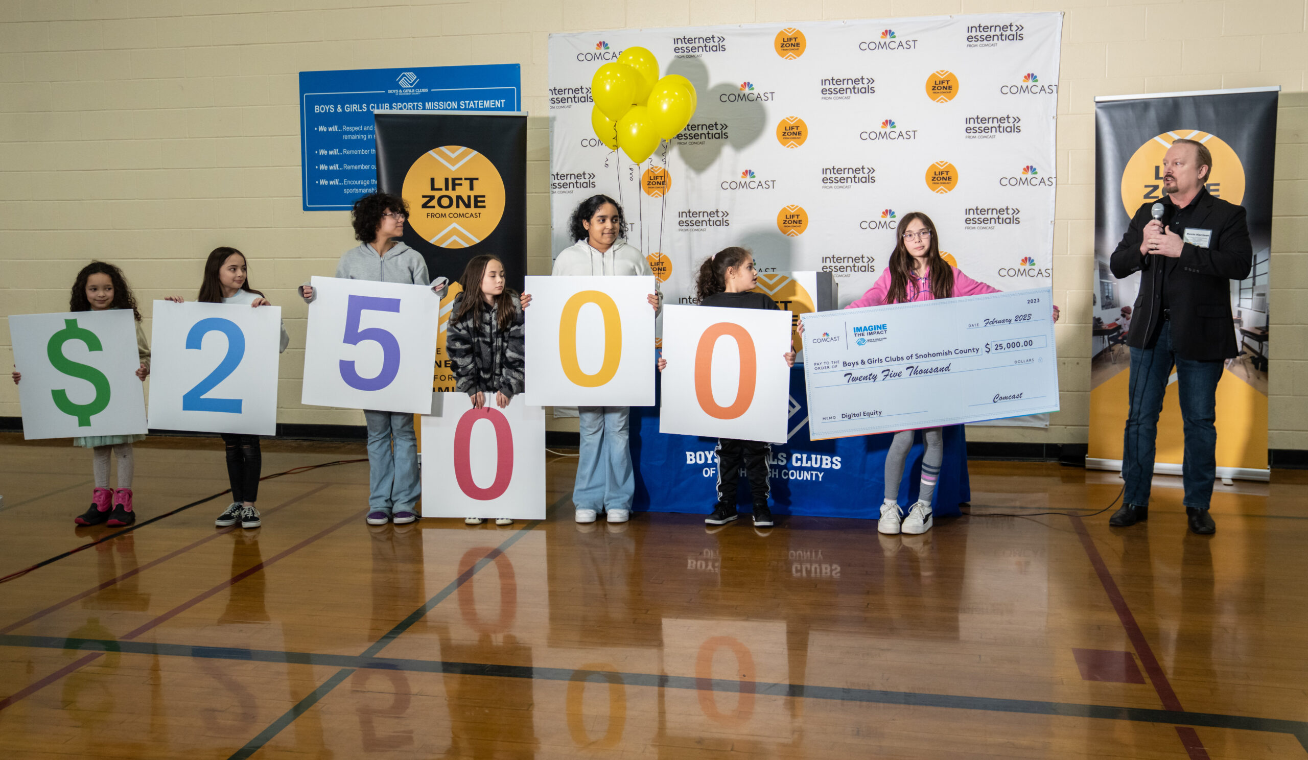 Children from the Boys & Girls Club Alderwood hold up signs of number values - 25,000 - in a gym. 