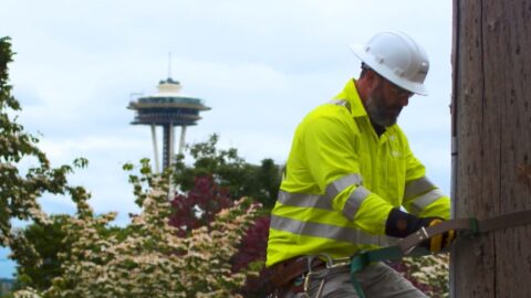 A Comcast technician works on a power pole with the Space Needle in the background.