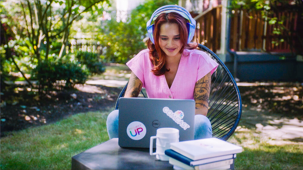 A photo of a young woman with red hair sitting outside at a laptop with headphones on.