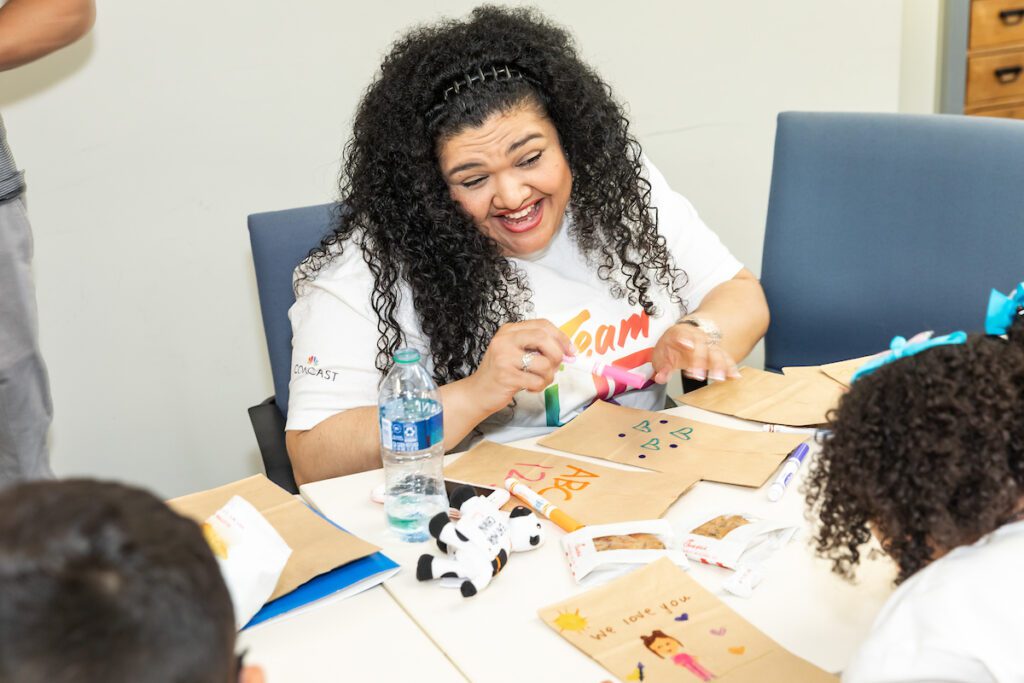 Woman with long curly hair smiling and chatting with child while decorating paper lunch bags