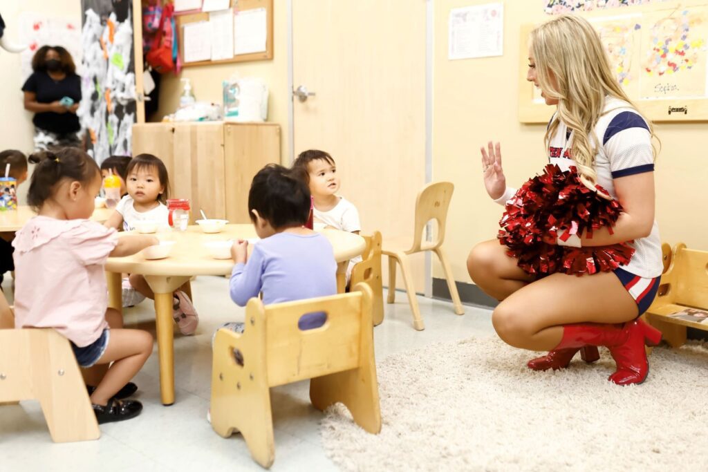 Cheerleader crouched low to speak to toddlers sitting at round wooden table