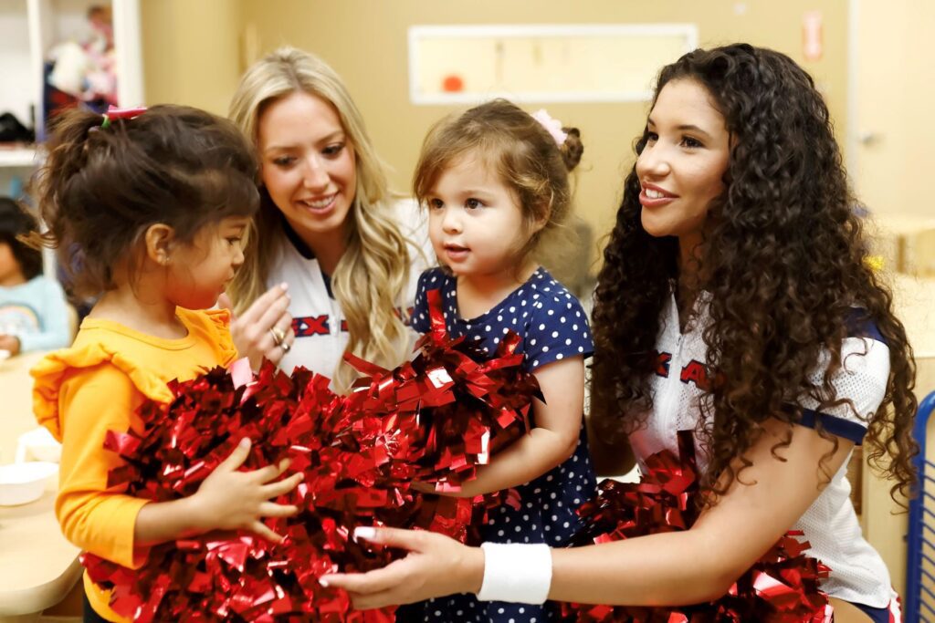 Two cheerleaders sitting with two young girls and sharing their red pom moms