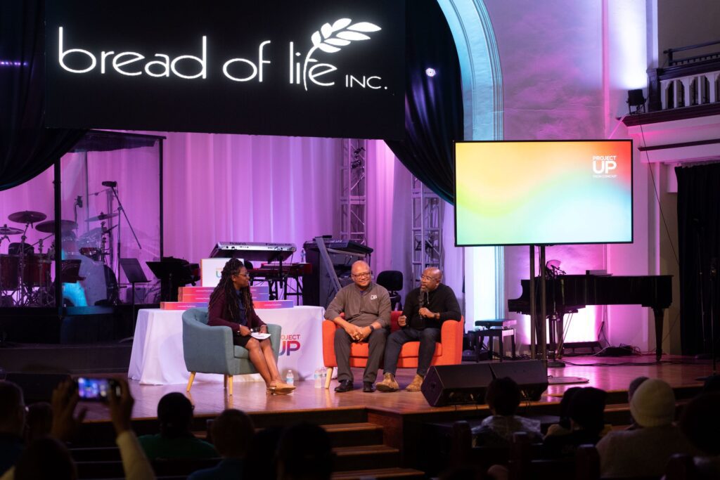 Three speakers sitting on orange sofa on event stage.