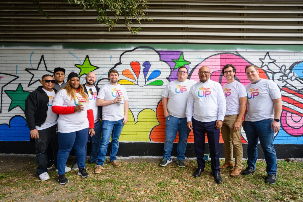 A group of Team Up volunteers posing by mural near painted Comcast logo peacock