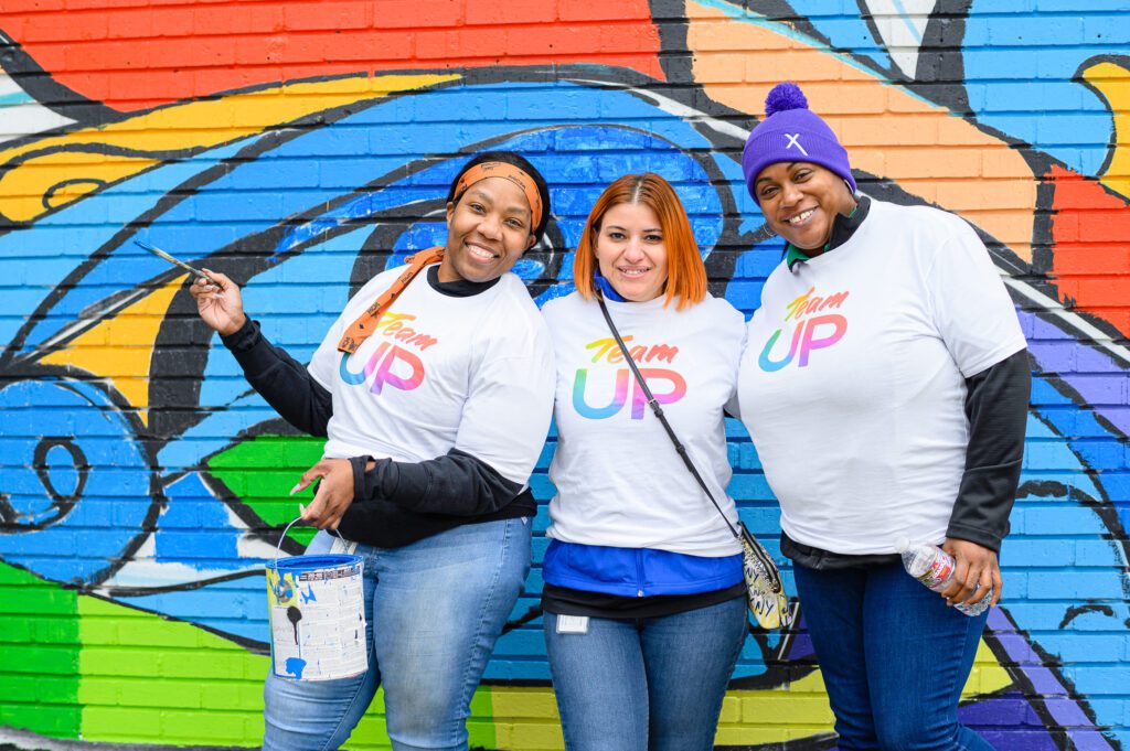 Three female volunteers smiling and posing for a picture near mural