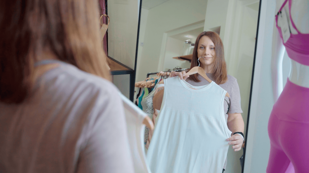 Woman looking in mirror holding up a hanger with a white tank top toward herself