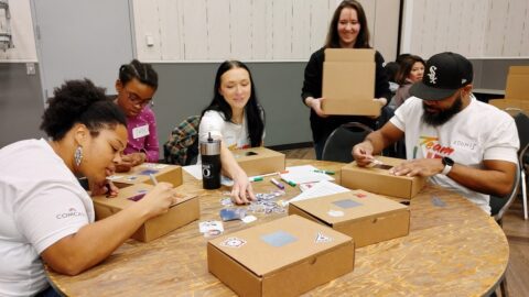 Five people work on cardboard boxes on a table.