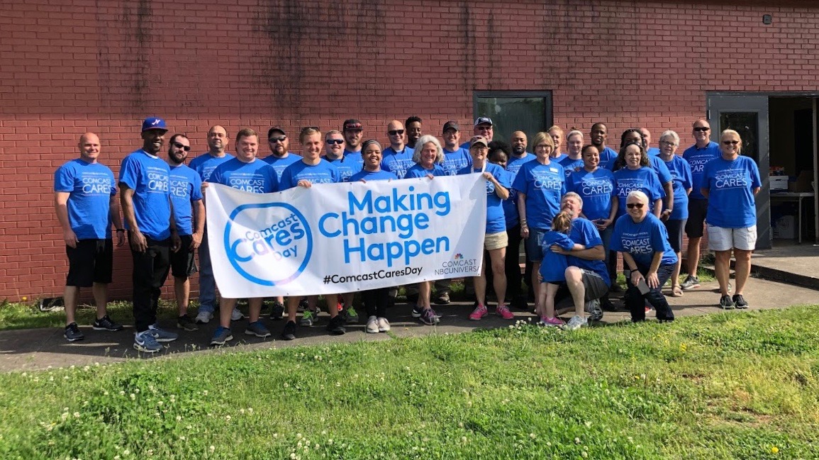 A large group of Comcast Cares Day volunteers hold a Making Change Happen banner.