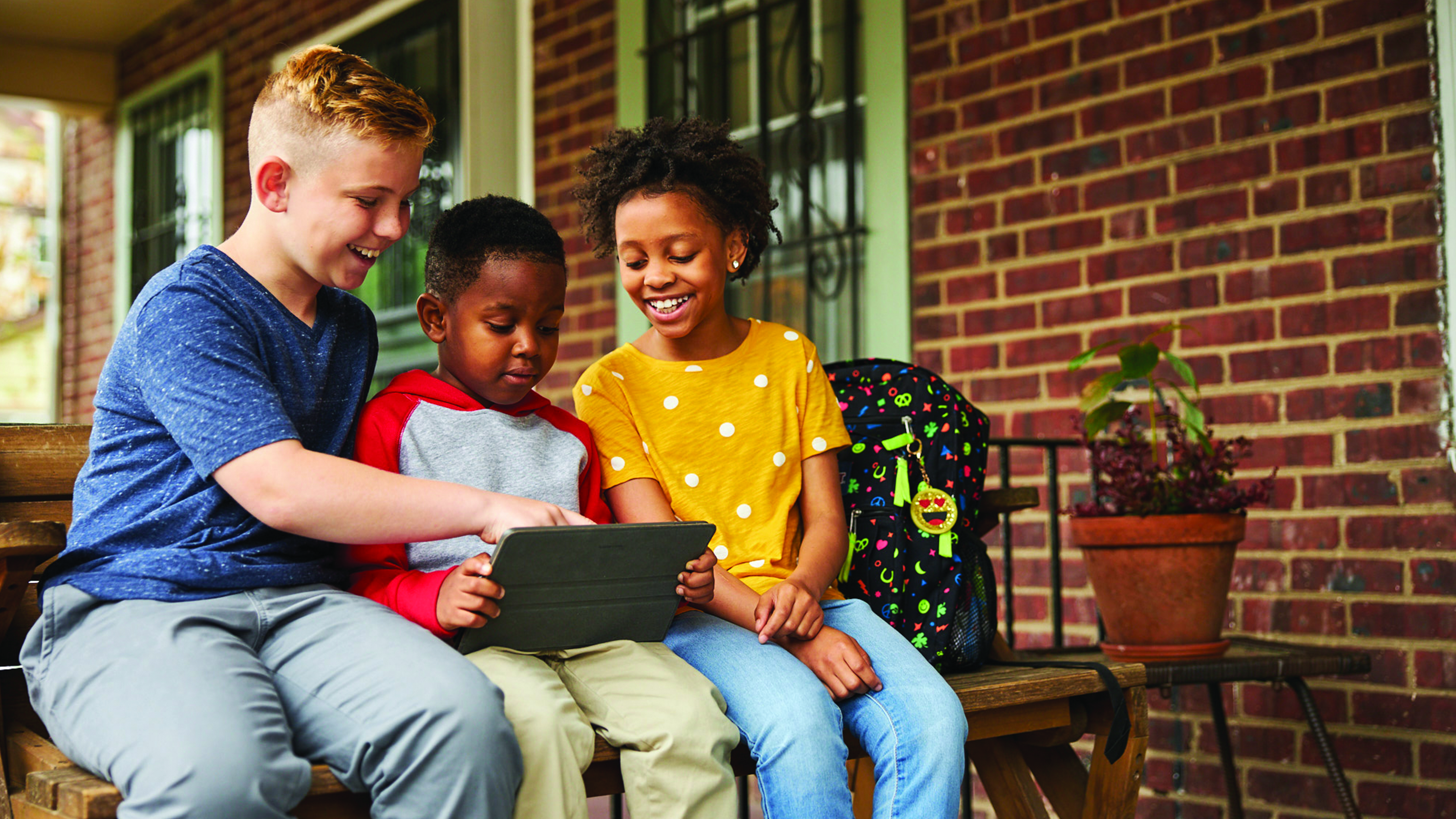 Three children use a tablet as they sit on a porch.