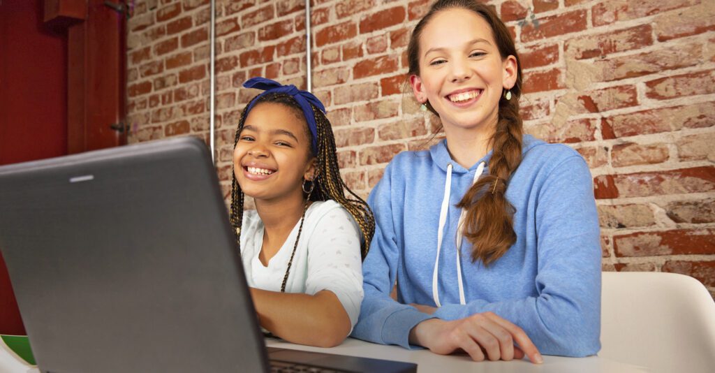 Two teen girls smile while using a laptop